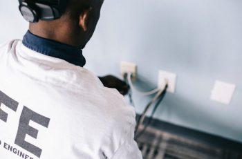 From above back view unrecognizable black field engineer wearing white shirt and protective hardhat sitting on floor and checking voltage in wall plugs